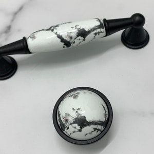 Op de afbeelding: Twee zwarte en witte keramische kastknoppen met een zwarte metalen basis. De grotere knop is een trekknop met een lange, rechthoekige vorm. De kleinere knop is rond. Beide knoppen hebben een zwart en wit abstract ontwerp.