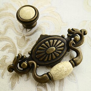 Könnte beinhalten: Antiker Bronze-Schrankgriff mit dekorativem Blumendesign und einem cremefarbenen Keramikknopf.
