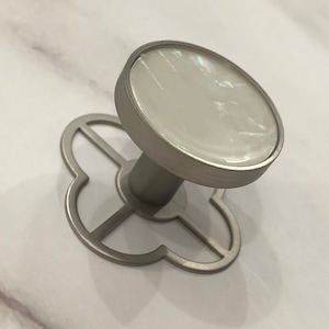 May include: A silver cabinet knob with a white mother-of-pearl center. The knob has a decorative, four-leaf clover design.