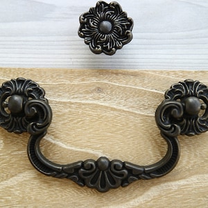 Könnte beinhalten: Ein Paar verzierte, antike Schrankgriffe mit einem dekorativen Blumenmuster. Die Griffe sind aus Metall und haben eine dunkelbraune Oberfläche.