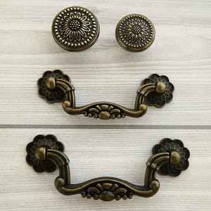 May include: Two antique brass drawer pulls with ornate floral designs. The pulls have a curved shape and are attached to a wooden surface.