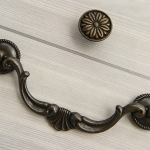 以下が含まれることがあります： 曲線的なデザインと装飾的なディテールが施された、装飾的なブロンズ製のキャビネットプル。プルは両端に丸いノブが付いています。