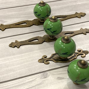 May include: Four green ceramic cabinet knobs with a leaf design and gold metal accents. The knobs are attached to ornate gold metal drawer pulls.