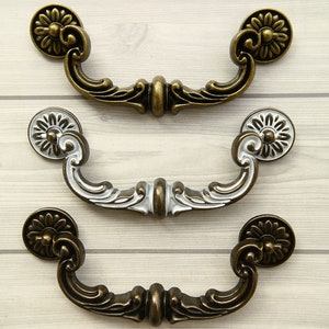 May include: Three ornate drawer pulls with a decorative floral design. The pulls are made of metal and have a distressed bronze finish. The top pull is bronze, the middle pull is white with bronze accents, and the bottom pull is bronze.