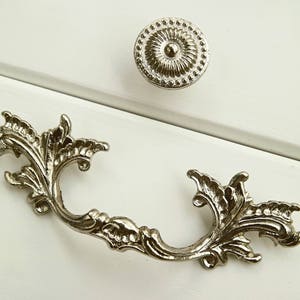 Puede incluir: Tirador de cajón adornado plateado con un diseño floral decorativo. El tirador tiene forma de 'C' con un asa curva.