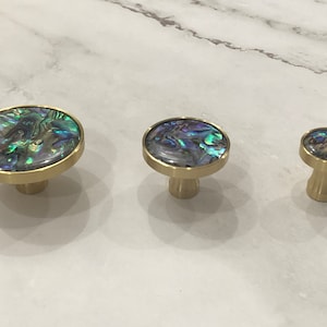 Könnte beinhalten: Drei Messing-Schrankknöpfe mit irisierenden Perlmutt-Einlagen. Die Knöpfe sind in verschiedenen Größen, wobei der größte im Vordergrund und der kleinste im Hintergrund ist.