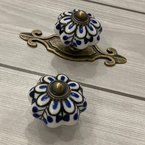 May include: Two blue and white ceramic drawer knobs with a floral design. The knobs have a brass finish and are attached to a decorative brass plate.
