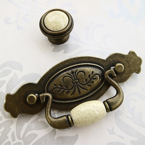 Könnte beinhalten: Antiker Messing-Schrankgriff mit einem ovalen Keramik-Knopf. Der Griff hat ein dekoratives Blumenmuster und der Knopf hat eine gesprenkelte cremefarbene Oberfläche.