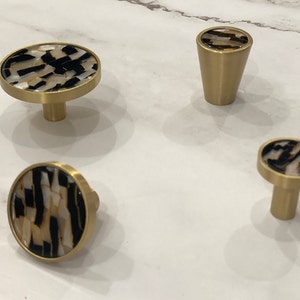 Puede incluir: Cuatro pomos de armario dorados con un diseño de patrón blanco y negro. Los pomos son redondos y tienen un pequeño vástago.