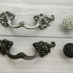 May include: Two sets of ornate drawer pulls with a floral design. One set is white and the other is bronze. Each set includes a pull and a knob.
