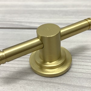 May include: A gold-toned metal towel hook with a round base and two arms extending out. The hook is designed to be mounted on a wall.