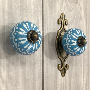 Puede incluir: Dos pomos de armario de cerámica azul con diseños florales blancos y detalles en latón antiguo. Los pomos están montados en una superficie de madera blanca.