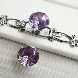 Puede incluir: Dos tiradores de cajón de cristal morado con base de metal plateado. El tirador más grande tiene un diseño decorativo con un motivo floral. El tirador más pequeño es un simple cristal redondo.