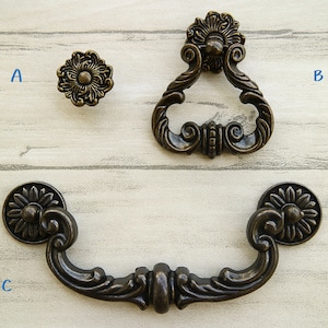 Pode incluir: Três puxadores de armário de estilo antigo em acabamento bronze. O primeiro puxador é um botão redondo com um design floral. O segundo puxador é um puxador de anel com um design floral. O terceiro puxador é um puxador em forma de D com um design floral.