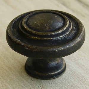 Könnte beinhalten: Ein runder, schwarzer Metall-Schrankknauf mit einem erhabenen, konzentrischen Kreisdesign. Der Knauf hat eine leicht gealterte, antike Oberfläche.