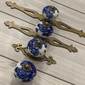 May include: Four blue and white porcelain cabinet knobs with gold metal accents and decorative backplates. The knobs feature a floral design.