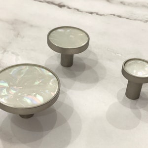 Può includere: Tre pomelli per armadi rotondi, bianchi e iridescenti con una base in metallo argentato.