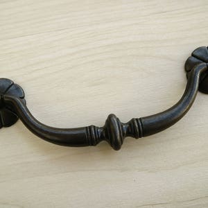 以下が含まれることがあります： 両端に装飾的な花柄が施された黒い金属製の引き出しの取っ手。取っ手は湾曲した形状で、中央が丸みを帯びています。