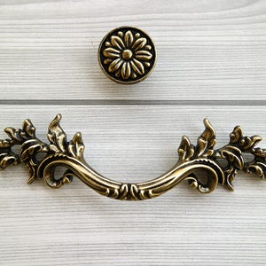 Może przedstawiać: Złoty, ozdobny uchwyt do szuflady z kwiatowym wzorem. Uchwyt ma kształt stylizowanego liścia z zakrzywionym uchwytem.