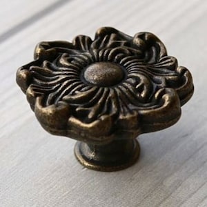 May include: A bronze-colored cabinet knob with a floral design. The knob has a round base and a raised, ornate top.