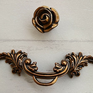 May include: A pair of ornate, gold-toned cabinet pulls. One pull is a rose-shaped knob, the other is a decorative, curved handle with leaf-like accents.