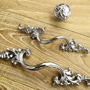 以下が含まれることがあります： 装飾的な花のデザインが施された、2つの装飾的な銀製の引き出しの取っ手。取っ手は、木製の表面に、それに合う銀製のノブと共に置かれています。