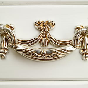 Puede incluir: Un tirador de cajón decorativo de plata y oro con un diseño de lazo y borla.