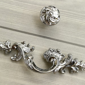 May include: A set of three ornate, silver-toned cabinet hardware pieces. The set includes a round knob with a floral design and a handle with a curved design and decorative flourishes. The hardware is designed for furniture.