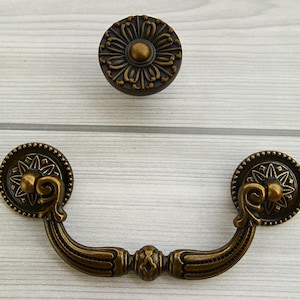 Könnte beinhalten: Ein Satz aus zwei antiken Messing-Schrankgriffen. Einer ist ein runder Knopf mit einem Blumenmuster und der andere ist ein dekorativer Griff mit einem gebogenen Griff.
