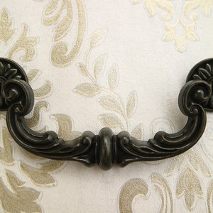 Puede incluir: Un tirador de cajón de metal negro con un diseño floral decorativo. El tirador tiene una forma curva y está diseñado para ser utilizado en armarios o cajones.