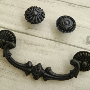 Könnte beinhalten: Schwarze Metall-Schrankbeschläge mit einem dekorativen Blumendesign. Die Beschläge umfassen einen Ziehgriff und zwei Knöpfe.