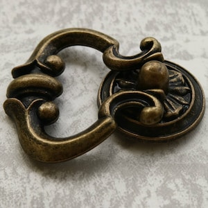 May include: A decorative antique gold-toned cabinet pull with a detailed floral design. The pull has a ring handle and a round base.
