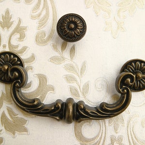 May include: Antique bronze cabinet hardware with a decorative floral design. The pull handle features a curved shape with ornate details. The knob is round with a raised floral pattern.