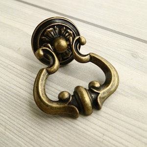 Puede incluir: Tirador de armario de latón antiguo con un asa de anillo decorativa y una base redonda y adornada.