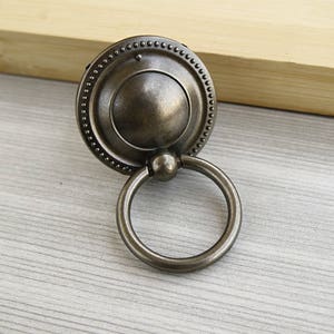 May include: A silver ring pull cabinet knob with a decorative, ornate, circular design. The knob has a ring pull handle.