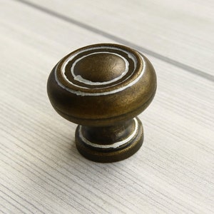 May include: A round, antique brass cabinet knob with white painted concentric circles. The knob has a slightly raised center and a smooth, polished finish.