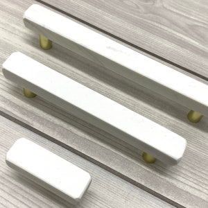 May include: Three white marble cabinet pulls with gold hardware. The pulls are rectangular in shape and have a smooth, polished finish.