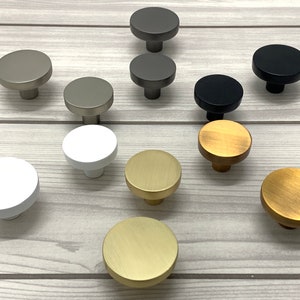 May include: Assortment of round cabinet knobs in various finishes, including brass, nickel, and black. The knobs are arranged on a light wood surface.
