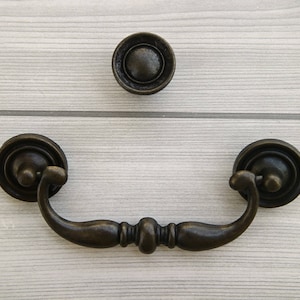 Könnte beinhalten: Ein bronzener Schrankgriff mit einem dekorativen Ring und einem gebogenen Griff.