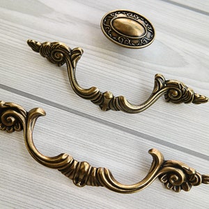 May include: Two ornate, antique-style, brass drawer pulls with a decorative floral design. The pulls are in the shape of a loop with a decorative scroll design on each side.