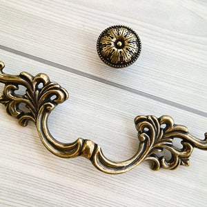 Może przedstawiać: Dekoracyjny mosiężny uchwyt do szuflady z kwiatowym wzorem. Uchwyt ma zakrzywiony kształt i okrągły guzik na końcu.