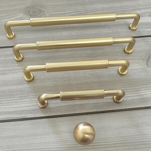 May include: A collection of four brushed gold cabinet pulls of varying lengths, each featuring a cylindrical center bar and two curved mounting posts, accompanied by a single matching round cabinet knob, all arranged in a vertical row.