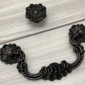 May include: Two antique-style cabinet hardware pulls in a dark bronze finish. One is a round knob with a floral design and the other is a decorative pull with a curved shape and ornate details.