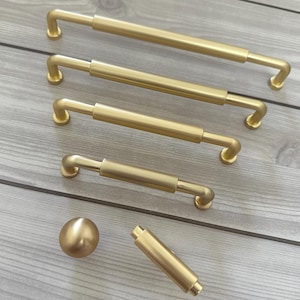May include: A collection of brushed gold cabinet hardware including four varying lengths of bar pulls, one spherical knob, and one cylindrical T-bar knob. Each piece features a smooth, matte metallic finish with clean lines and simple geometric shapes.