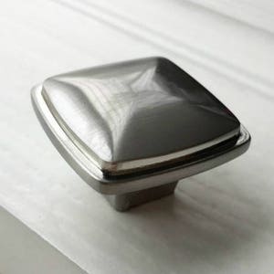 May include: A square-shaped cabinet knob with a brushed nickel finish. The knob has a raised, beveled edge and a smooth, flat surface.