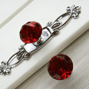 Peut inclure: Un bouton de cabinet en cristal rouge avec une base ornée argentée. Le bouton est fixé à une surface blanche.