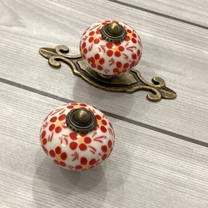 以下が含まれることがあります： 金色のアクセントが付いた赤と白のセラミック製の引き出しノブが2つ。ノブには花柄が施されています。