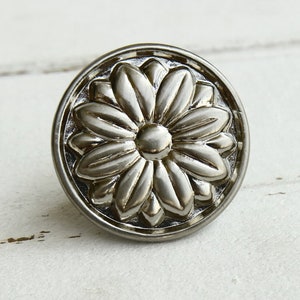 Puede incluir: Pomo de armario de metal plateado con un diseño floral en relieve. El pomo tiene forma circular y un acabado liso y brillante.