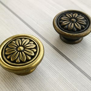 Könnte beinhalten: Zwei antike goldfarbene Schrankknöpfe mit Blumenmuster. Die Knöpfe haben ein erhabenes, kreisförmiges Design mit einer Blume in der Mitte.