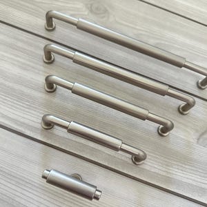 May include: A set of five brushed nickel cabinet pulls arranged by size from longest to shortest. Each pull features a cylindrical center bar with rounded ends and two small circular mounting bases that attach to the cabinet surface.